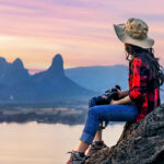 Woman enjoying a solo travel experience while sitting on a rocky viewpoint at sunset, overlooking mountains and a river.