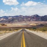 A straight desert highway leads into multicolored mountains under a blue sky with scattered clouds, capturing the essence of Route 66—one of the most iconic road trips in the USA.
