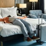 Exhausted traveler lying face-up on a hotel bed with suitcase nearby, in a cozy modern yet cheap hotel room.